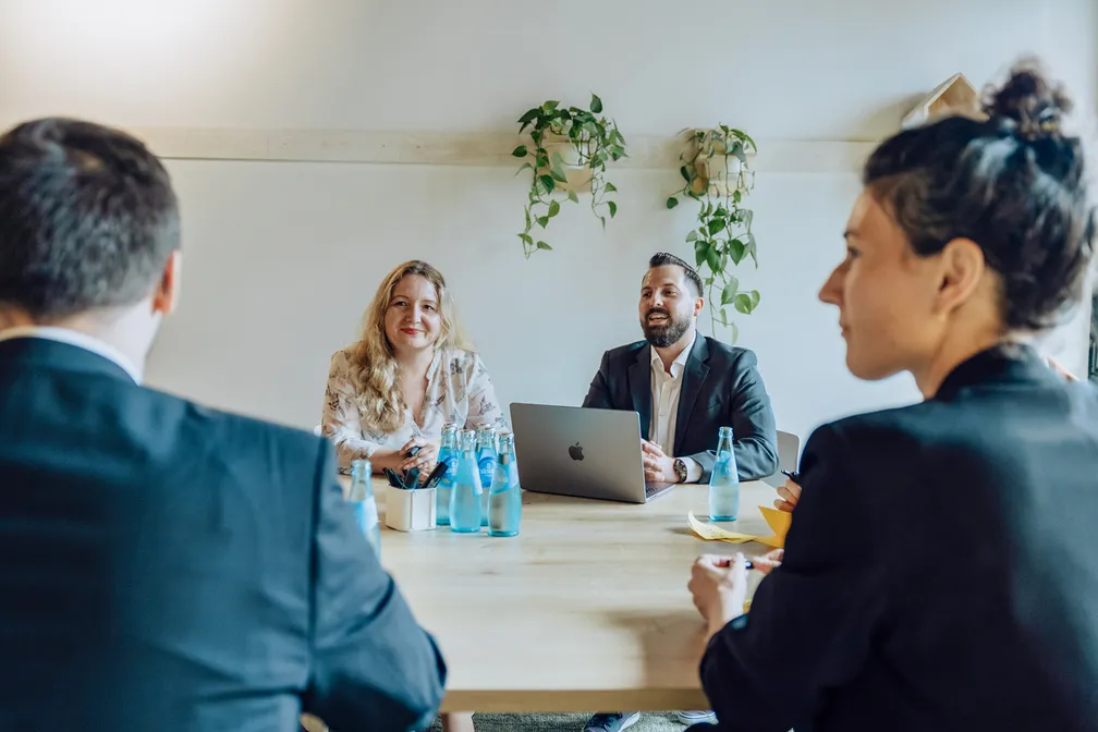 A group of four people in a business meeting, sitting around a table with laptops and water bottles, in a room with plants on the wall.