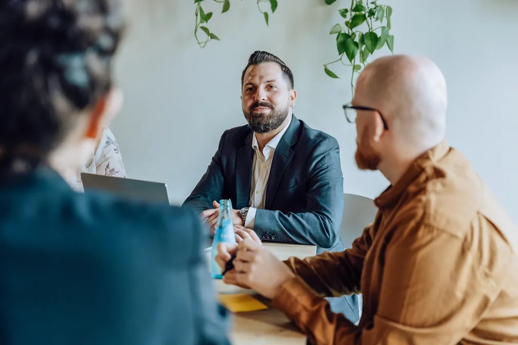 A group of people in a meeting room, with a man in a suit smiling and others listening attentively. There are laptops and a bottle on the table, and plants hanging in the background.