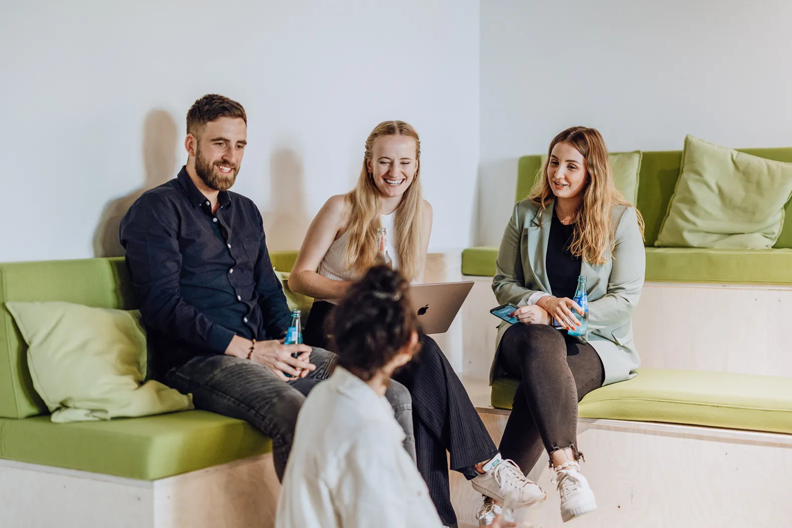 A group of four people sitting on green cushioned benches, engaged in a friendly conversation. One person is holding a laptop, while others have drinks in their hands. They are smiling and appear relaxed in a casual setting.