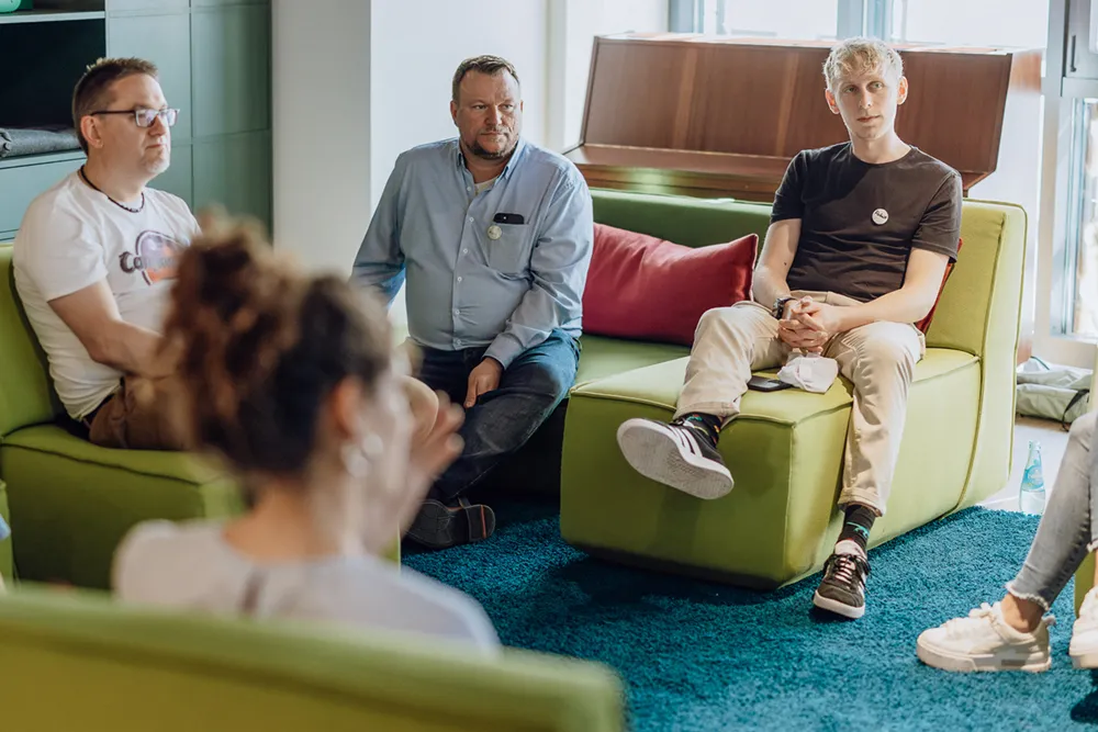 A group of people sitting on green couches in a casual meeting setting, with a piano in the background.