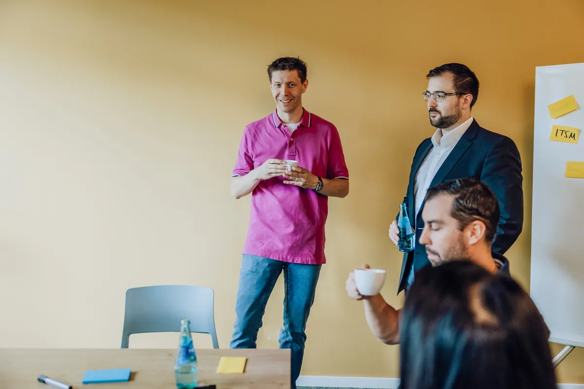 A group of people standing and sitting in a meeting room with a yellow wall, holding drinks and discussing near a whiteboard with sticky notes.
