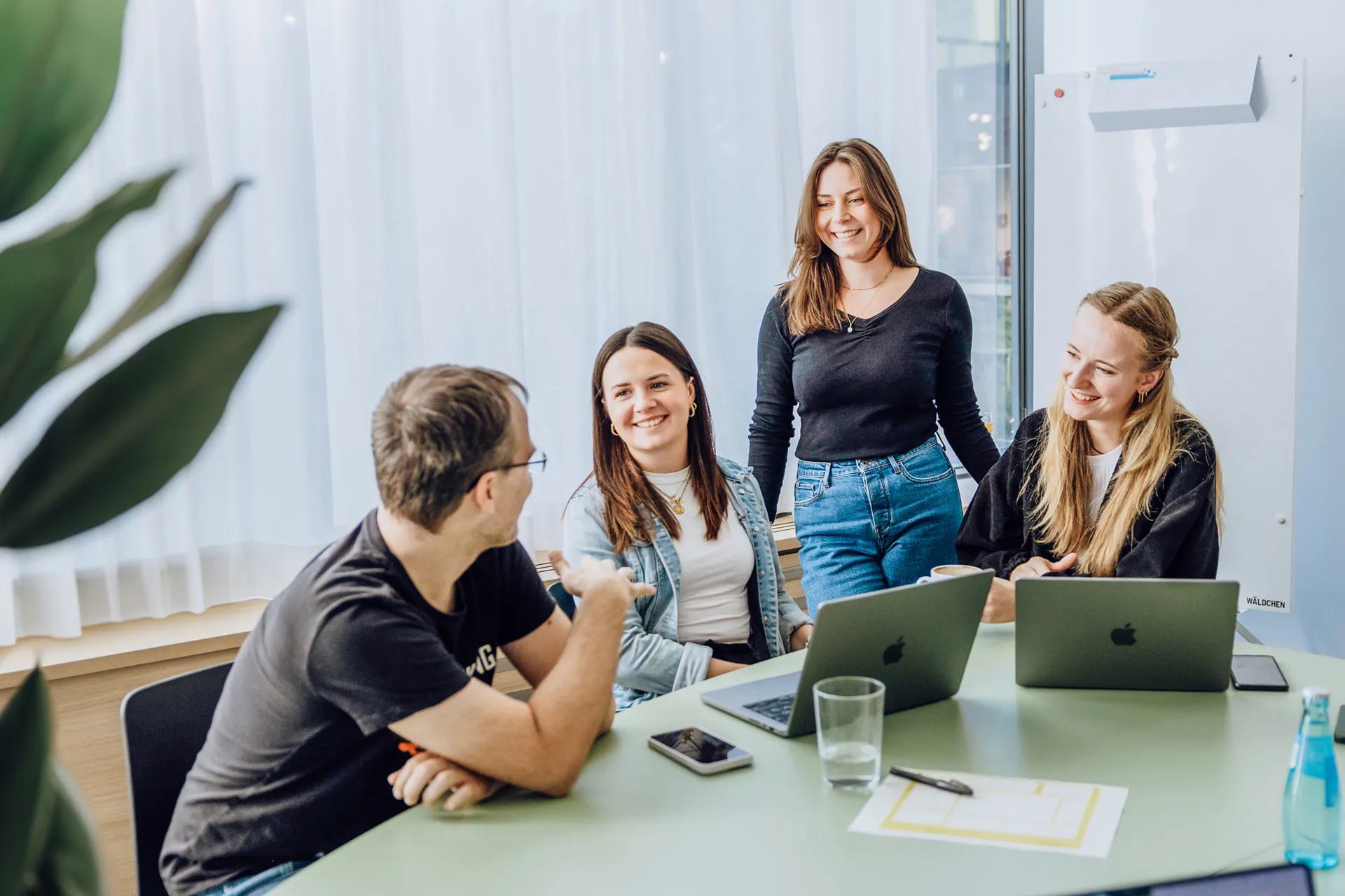 A group of four people sitting and standing around a table with laptops, smiling and engaging in a casual discussion in a bright office setting.