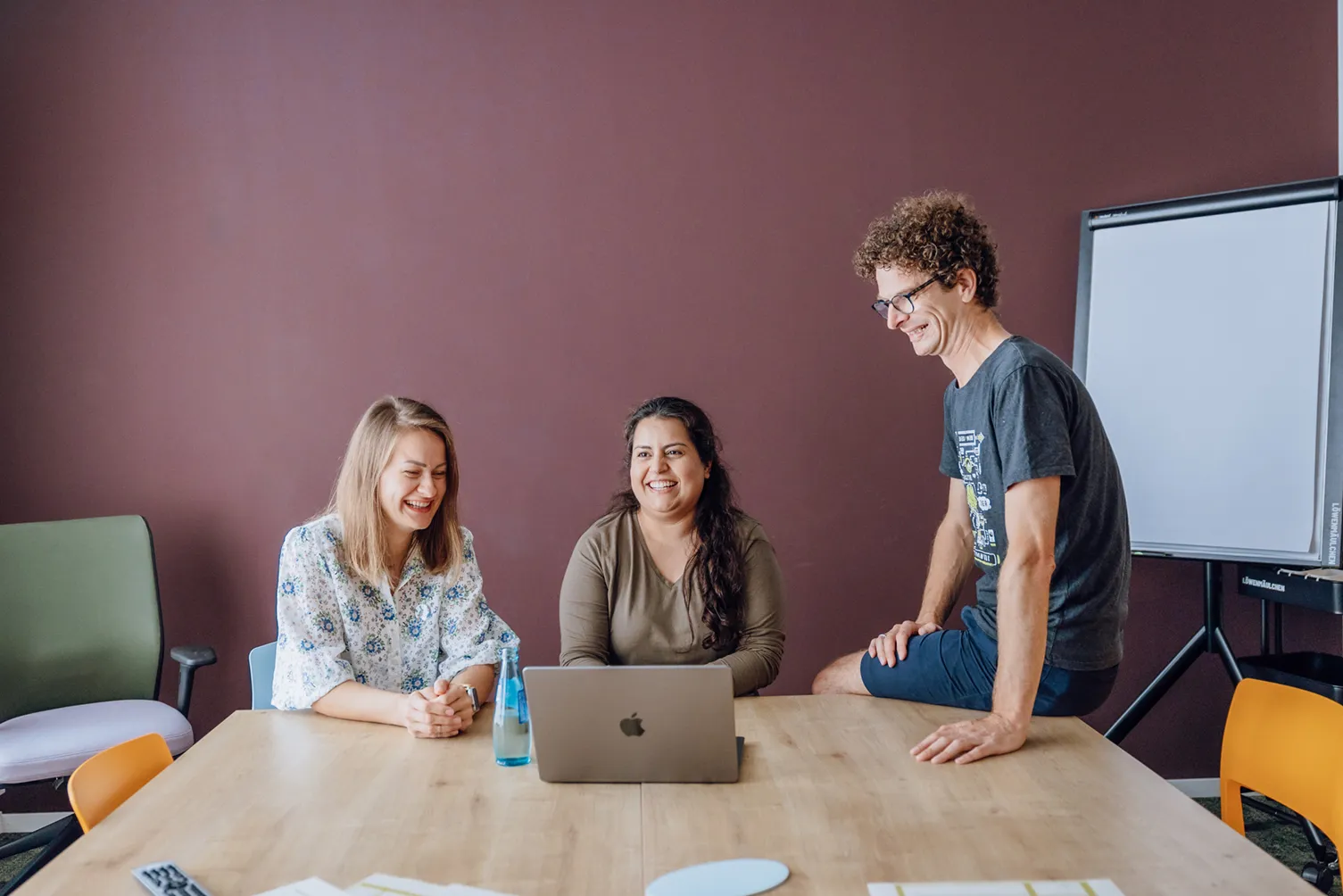 Three people smiling and sitting around a table with a laptop, in a casual office setting.