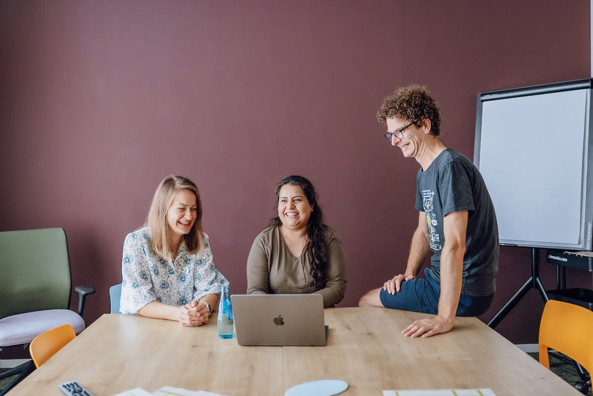 Three people smiling and sitting around a table with a laptop, in a casual office setting.
