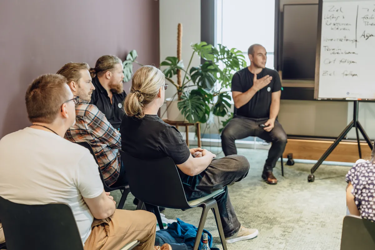 A group of people sitting in chairs attentively listening to a speaker in a casual office setting. The speaker is gesturing towards a flip chart with notes. There are plants in the background.