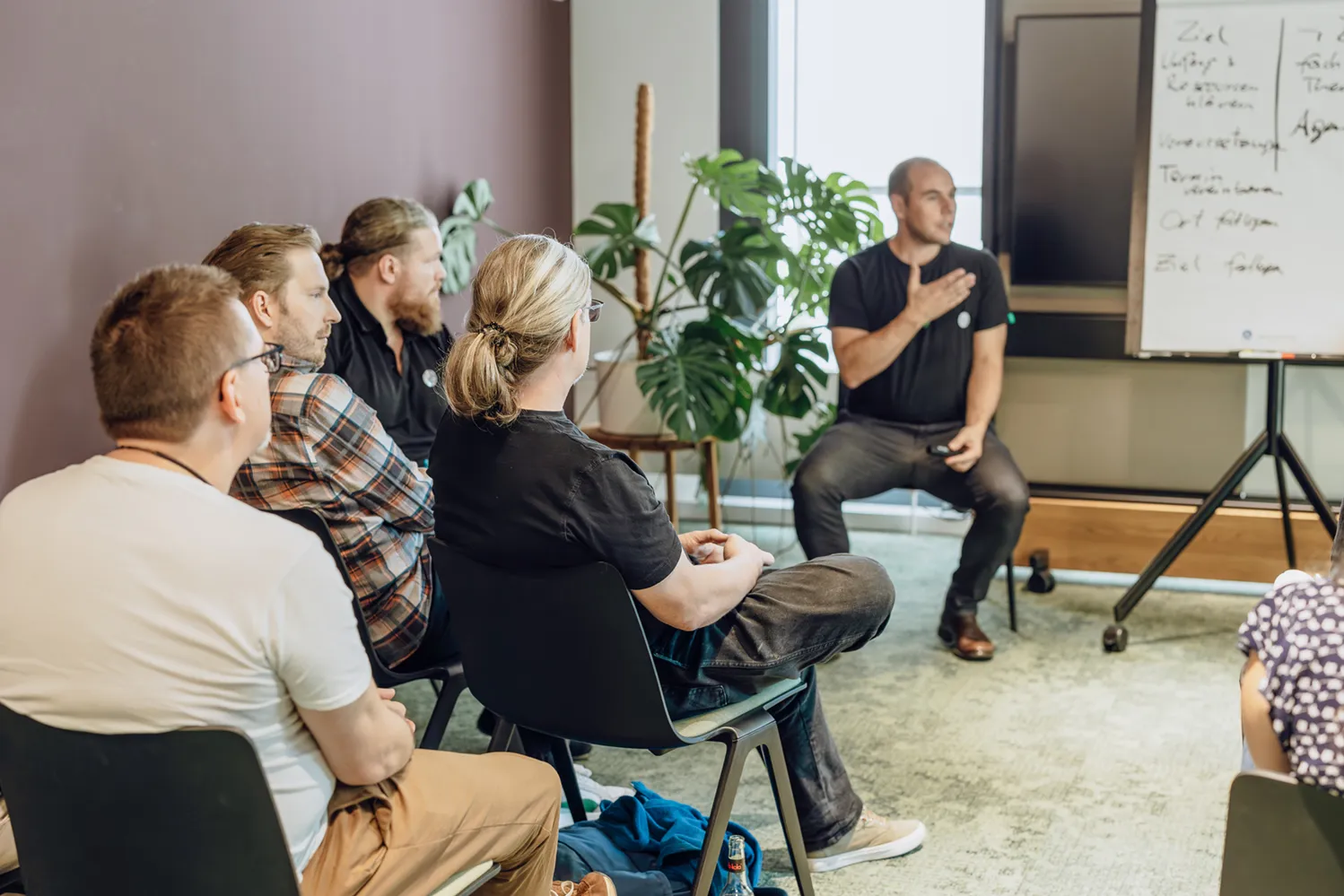 A group of people sitting in chairs attentively listening to a speaker in a casual office setting. The speaker is gesturing towards a flip chart with notes. There are plants in the background.