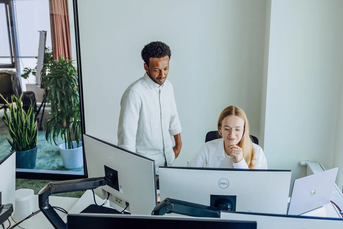 Two colleagues working together at a desk with multiple computer monitors in an office setting. One person is standing and the other is seated, both focused on the screen.