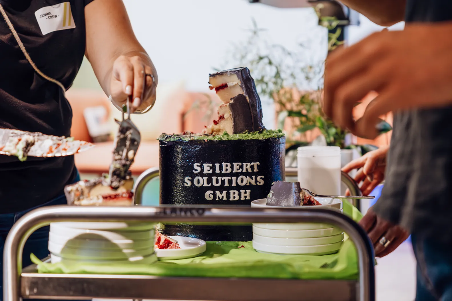 A person cutting a cake with the text 'Seibert Solutions GmbH' on it, placed on a table with plates and utensils.