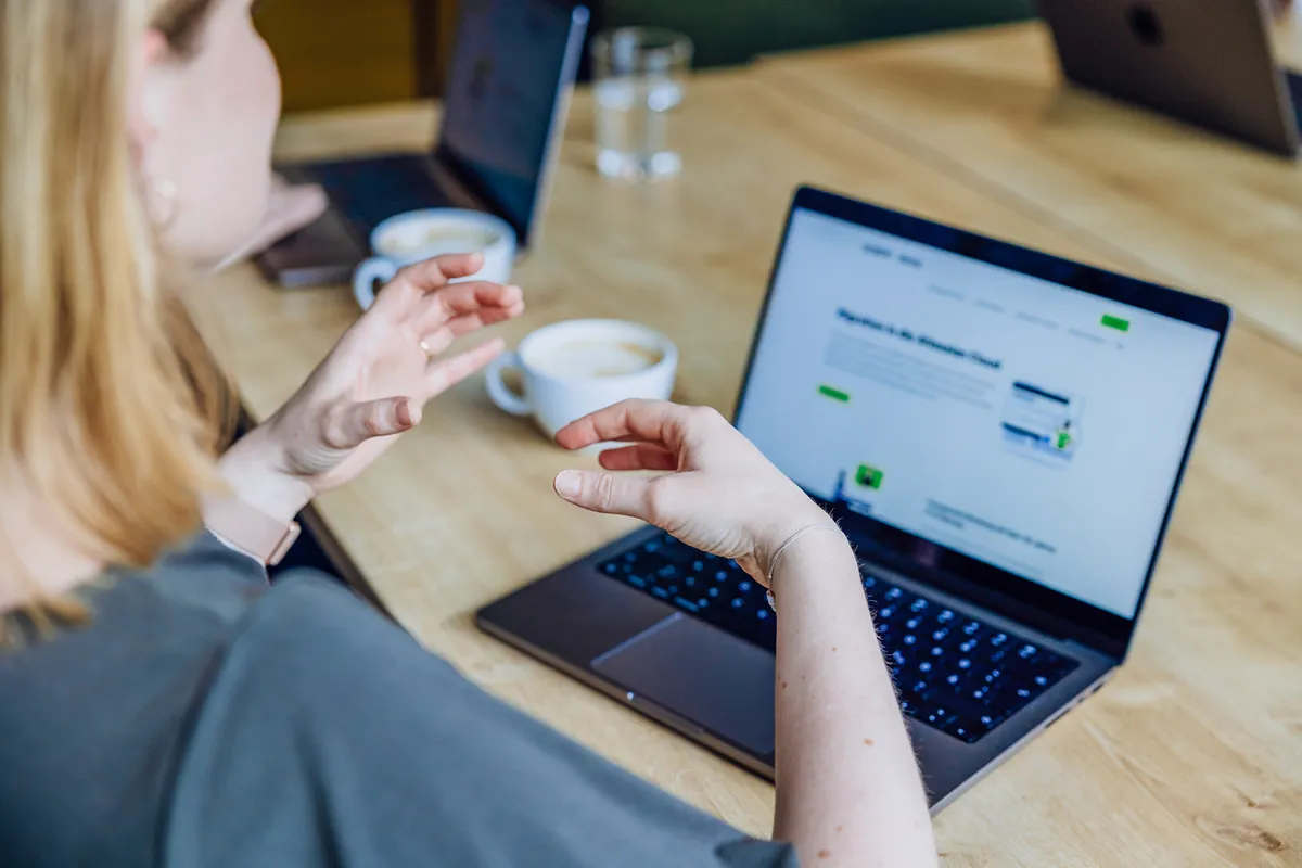 Person gesturing while discussing content on a laptop screen with coffee cups on the table.
