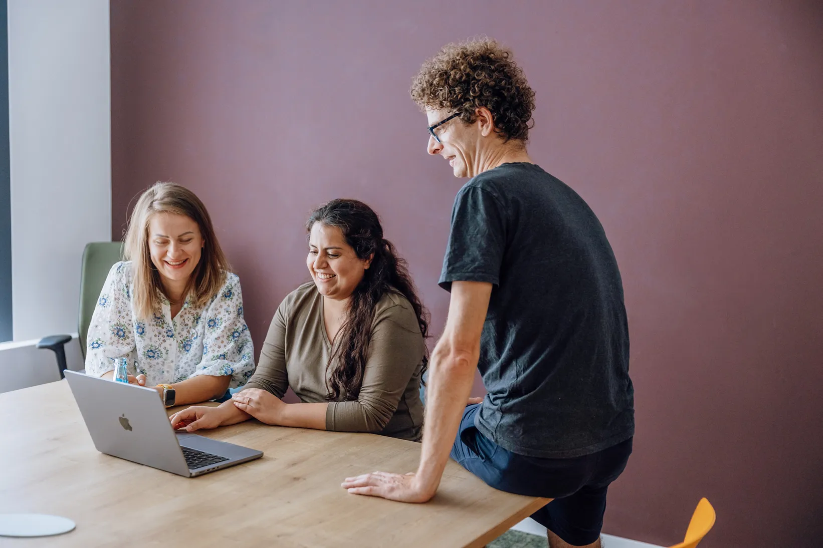 Three people gathered around a laptop on a wooden table, smiling and engaged in conversation.