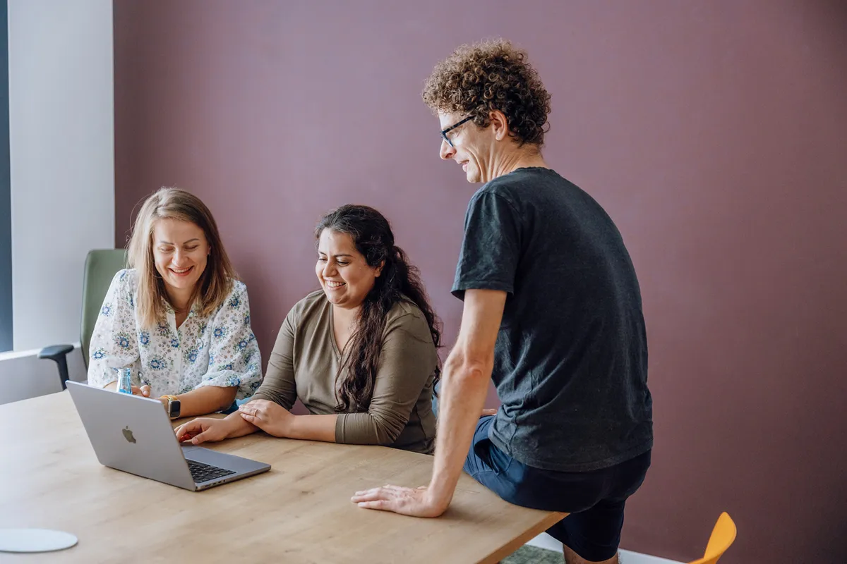 Three people gathered around a laptop on a wooden table, smiling and engaged in conversation.