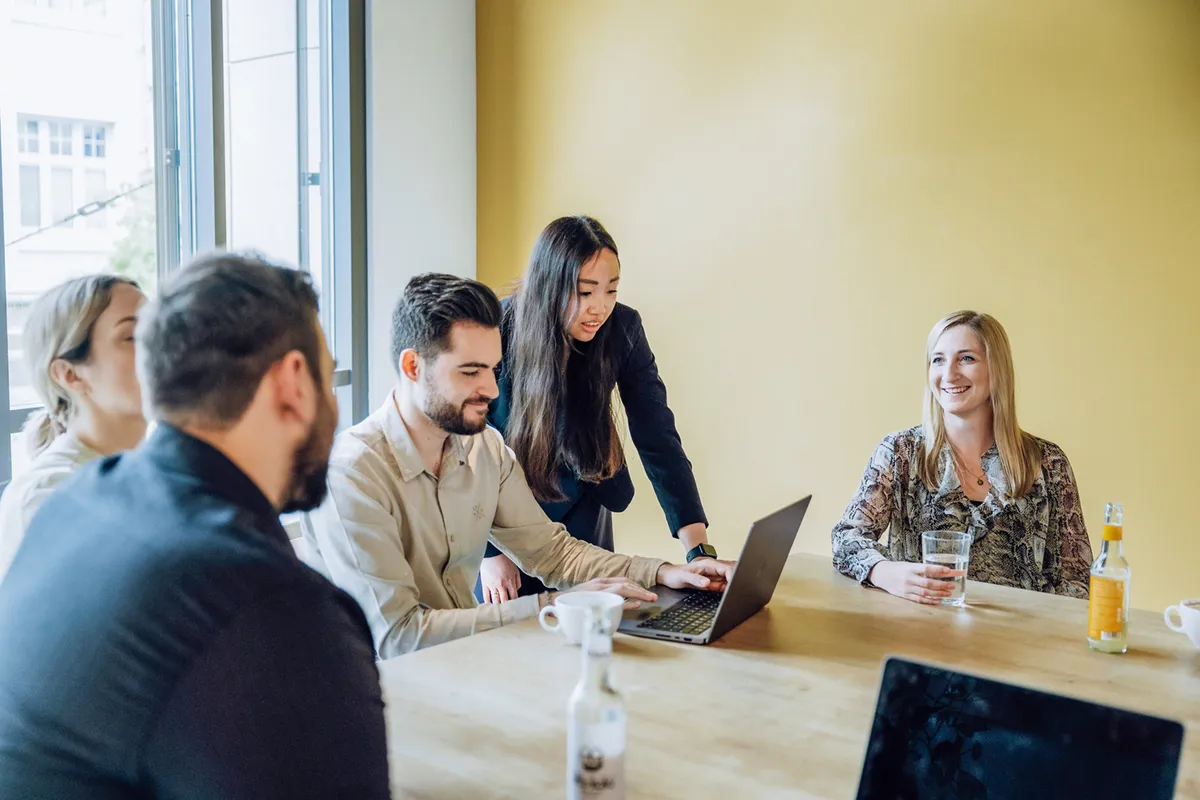 A group of people sitting around a table with laptops, engaged in a discussion. One person is standing and pointing at a laptop screen, while others are seated, smiling and holding drinks. The setting is a bright room with a yellow wall.