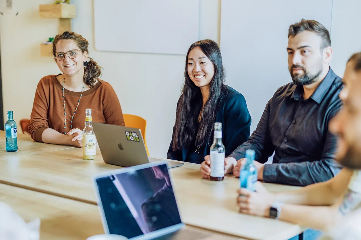 A group of people sitting around a table with laptops and drinks, engaged in a meeting or discussion.