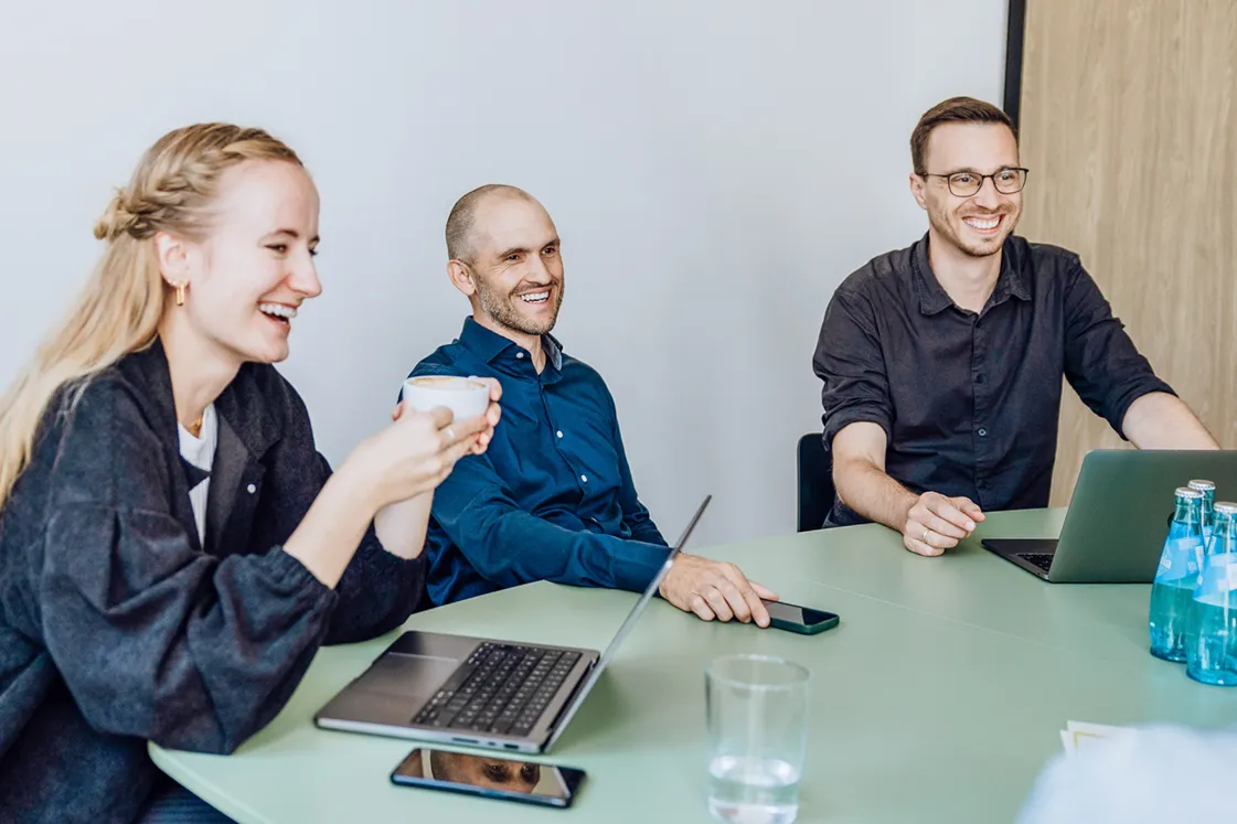 Three people sitting at a table, smiling and engaged in conversation. One person is holding a cup, and two laptops are visible on the table.