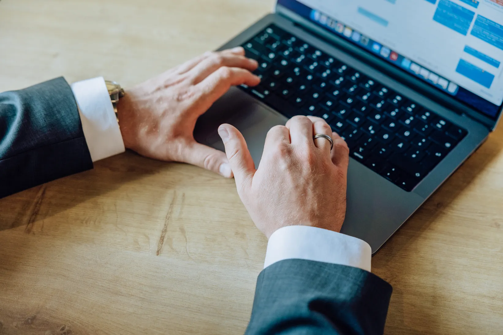 Close-up of hands typing on a laptop keyboard, wearing a suit and ring.