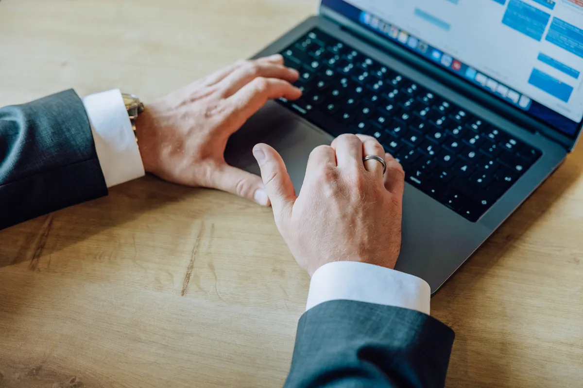 Close-up of hands typing on a laptop keyboard, wearing a suit and ring.