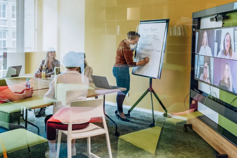 A person writing on a flipchart in a meeting room with several people sitting at a table and a video conference screen showing remote participants.