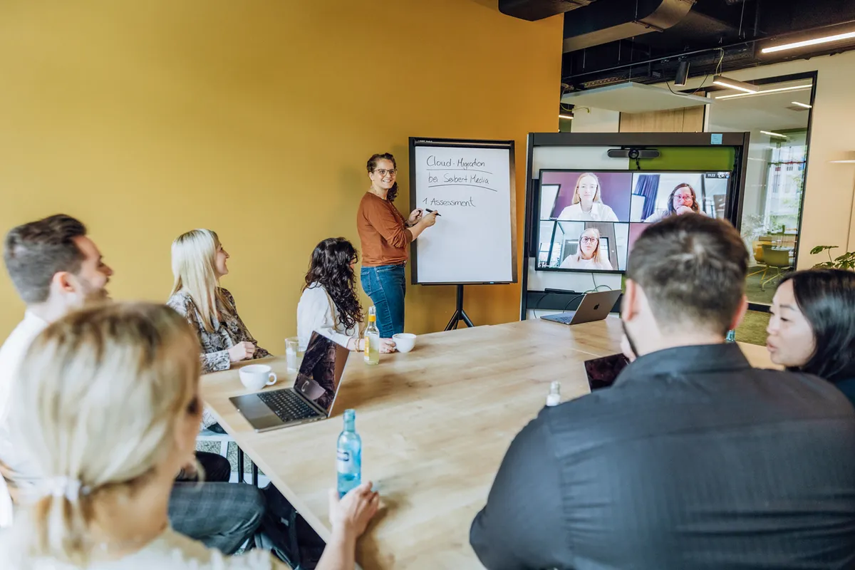 A group of people in a meeting room with a yellow wall, watching a presentation on a flipchart. A woman is standing and pointing at the flipchart, while others are seated around a table with laptops. A video conference screen shows remote participants.