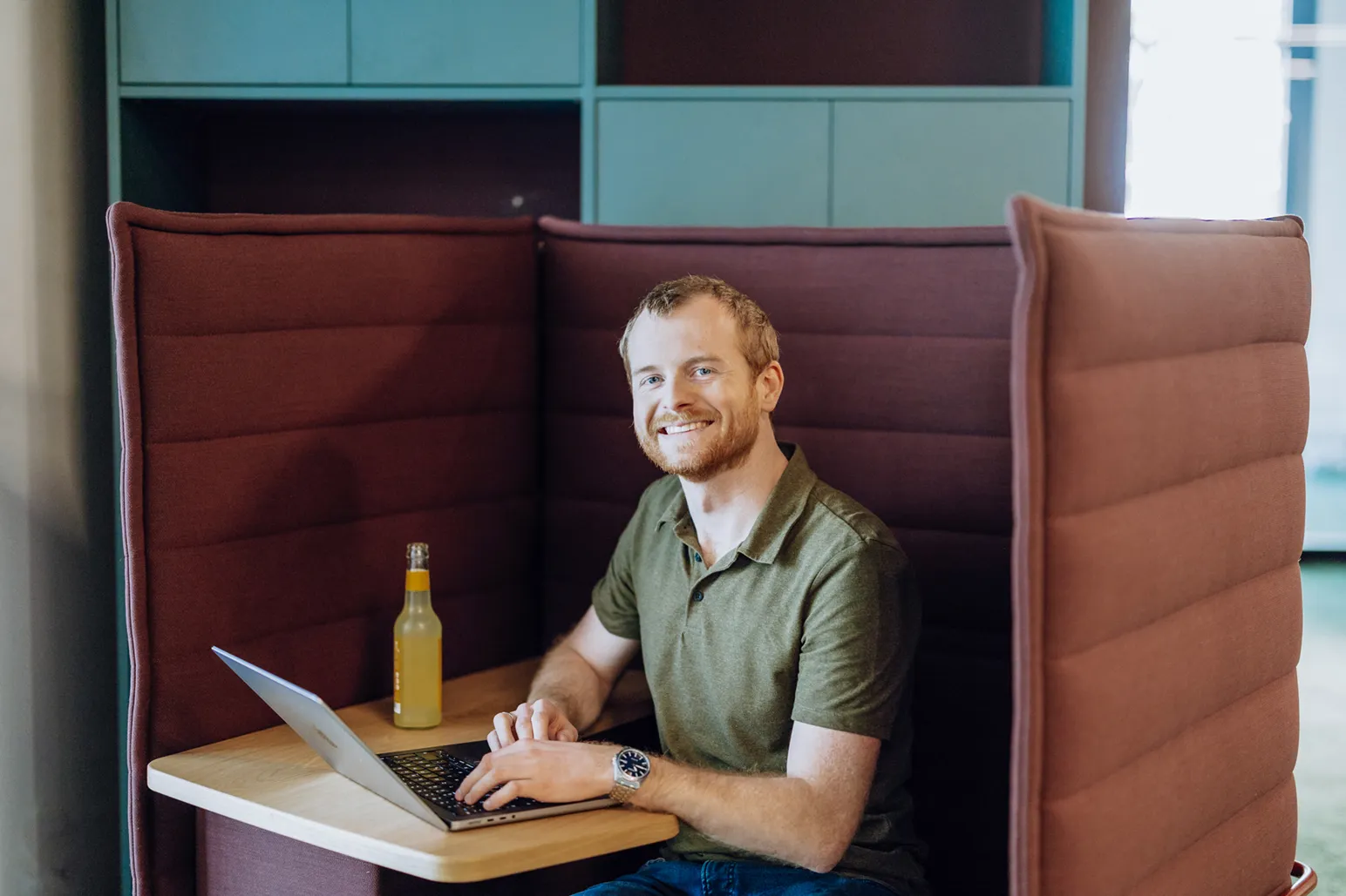 A man sitting in a booth with a laptop and a bottle, smiling at the camera.