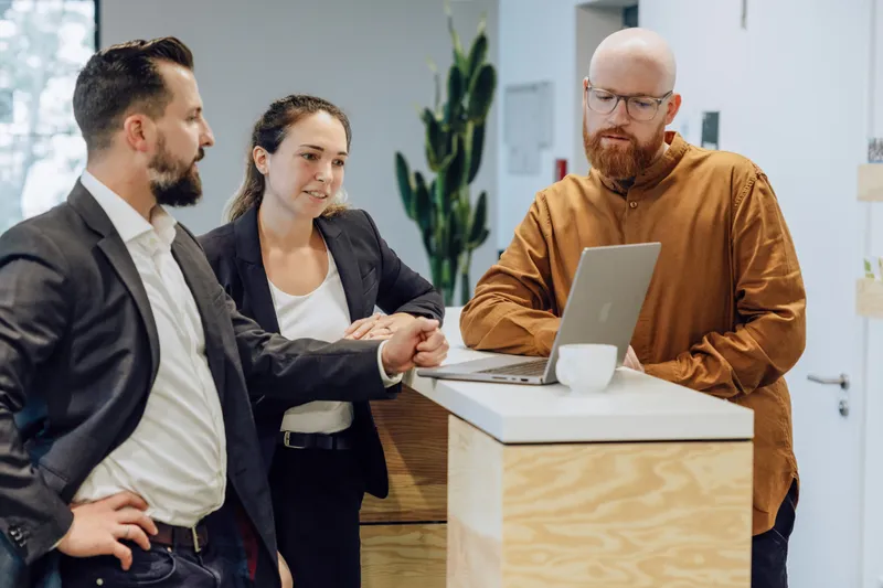Three people in business attire having a discussion around a laptop on a wooden counter in an office setting.