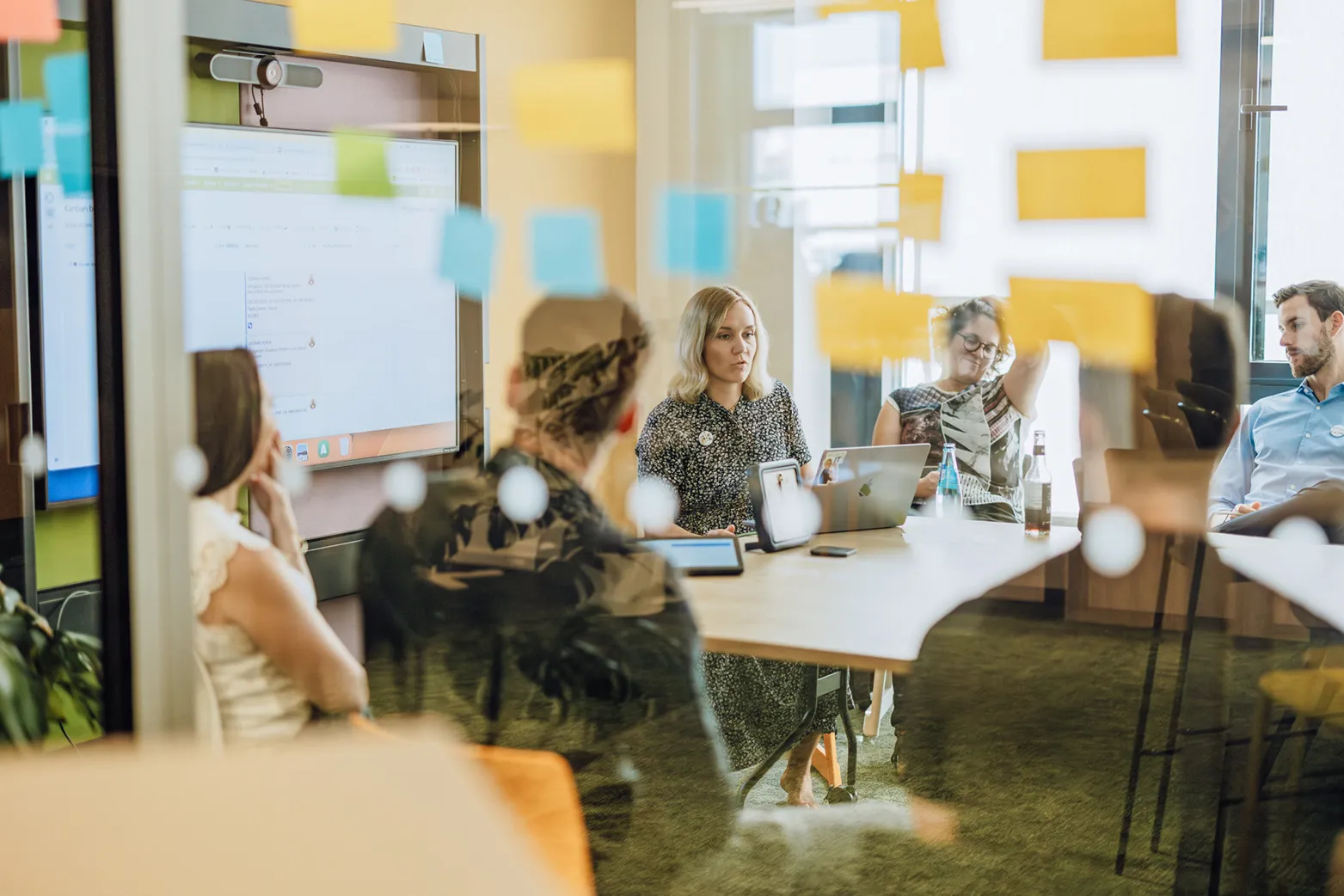Office meeting with people sitting around a table, visible through a glass wall with sticky notes.