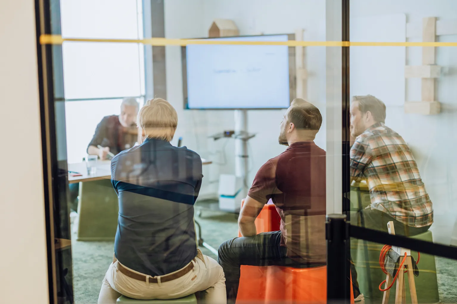 A group of people sitting in a modern office room, attentively watching a presentation on a large screen. The room is visible through a glass wall.
