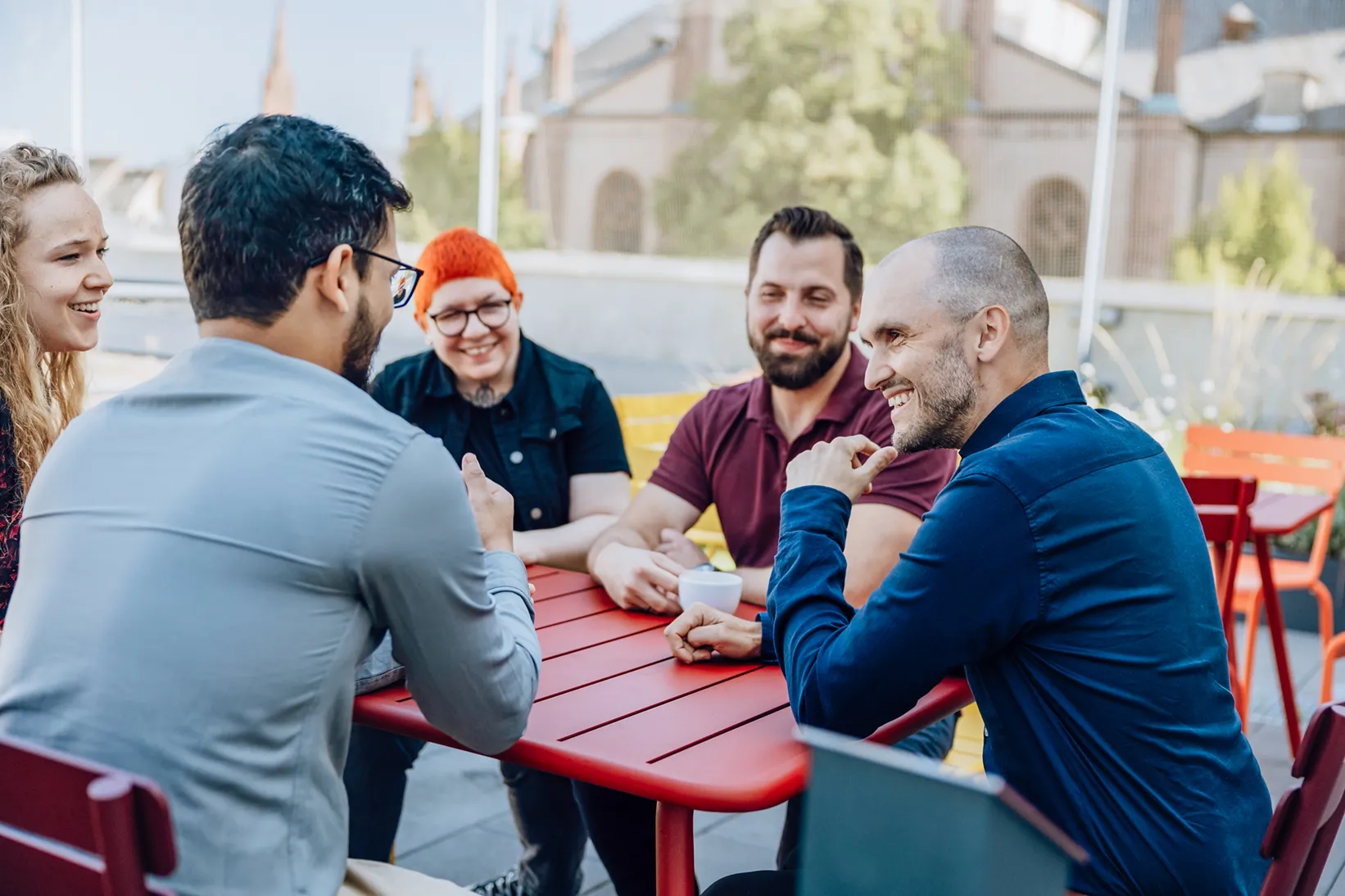 A group of five people sitting around a red table outdoors, engaged in a lively conversation. They are smiling and appear to be enjoying each other's company. The background shows a blurred view of buildings and greenery.