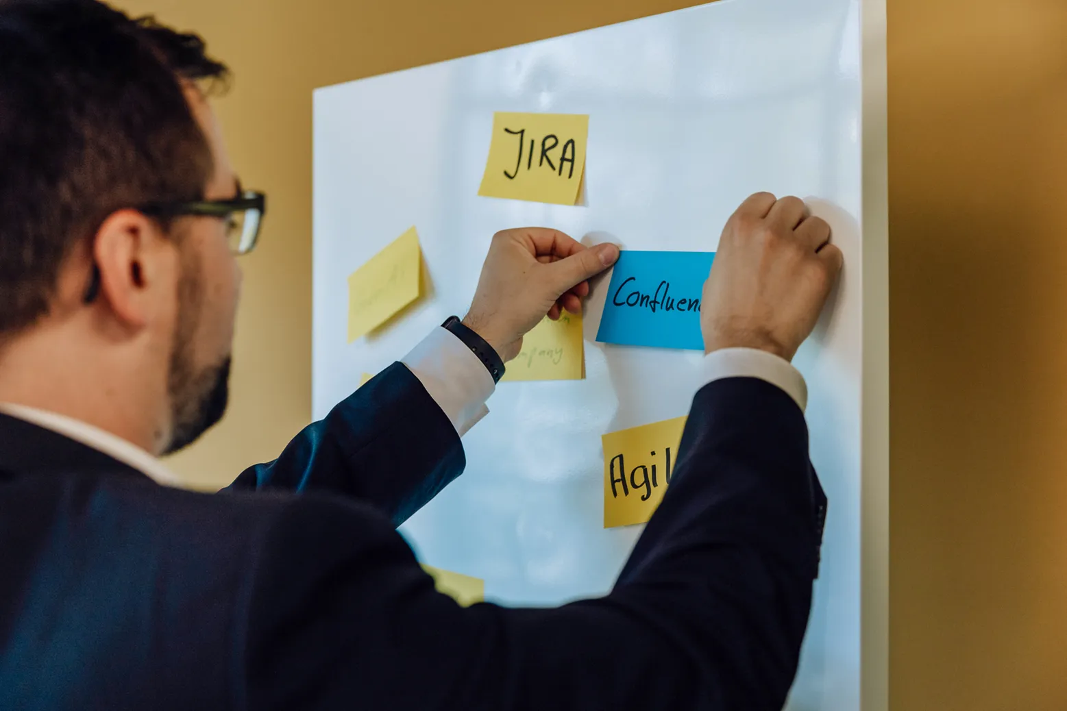 Person placing sticky notes on a whiteboard with words like 'JIRA' and 'Confluence'.