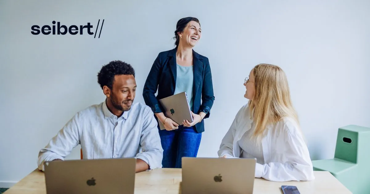 Three individuals, two seated at laptops and one standing, laughing and interacting in an office. The seibert// logo is in the top left.