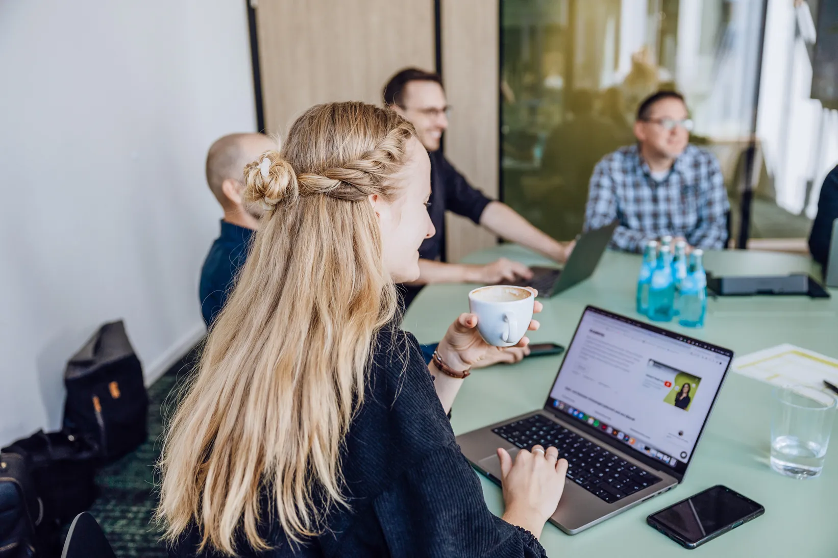 A woman with long blonde hair holding a coffee cup while using a laptop in a meeting room with colleagues.
