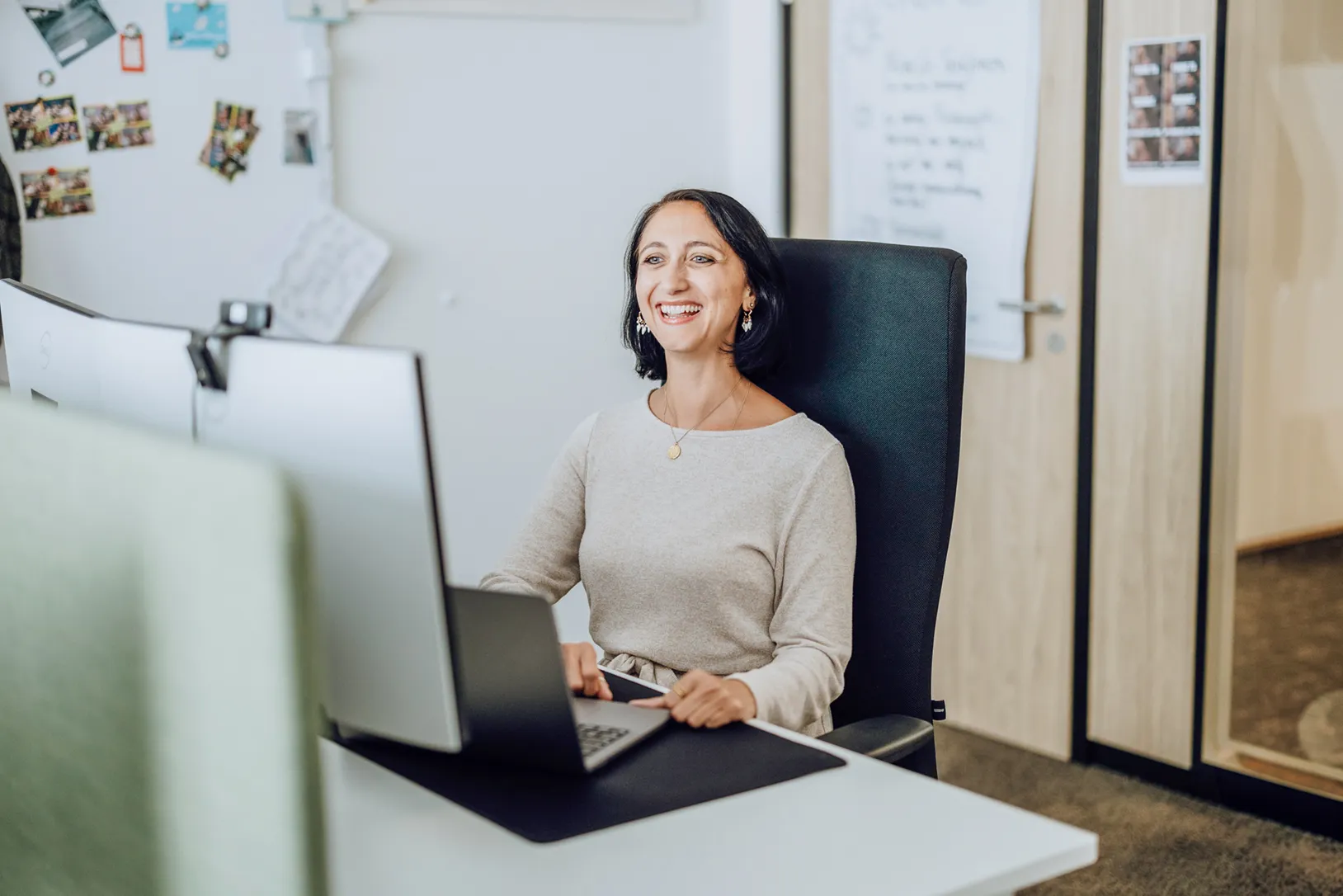 Woman smiling while working at a desk with a laptop and dual monitors in an office setting.